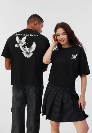 Sort bomulds t-shirt med et hvidt grafisk motiv af flyvende fugle og teksten "Kærlighed og Fred" på bagsiden; tætsiddende, kortærmet design.