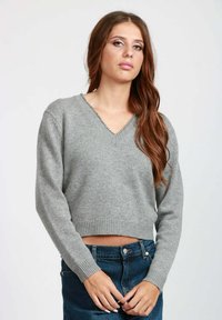 Giovane donna con lunghi capelli castani, che indossa un maglione grigio a V corta e jeans di denim blu, in piedi contro uno sfondo neutro.