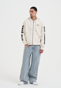 Felpa beige con zip e cappuccio regolabile, testo nero sulle maniche. Abbinata a jeans a palazzo blu chiaro e sneakers bianche.
