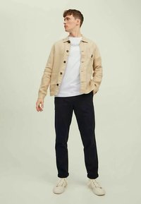 Béžna jakna s gumbi čez belo majico, kombinirana z mornarsko modrimi hlačami in belimi supergami, ustvarja priložnostni večplastni videz.