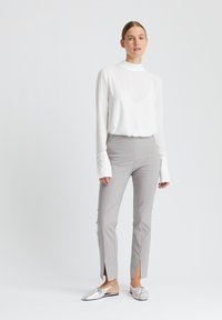 Weißes Langarmshirt mit hohem Kragen und Glockenärmeln; graue, schmal geschnittene Hose mit Seitenschlitzen; glänzende silberne Slipper.