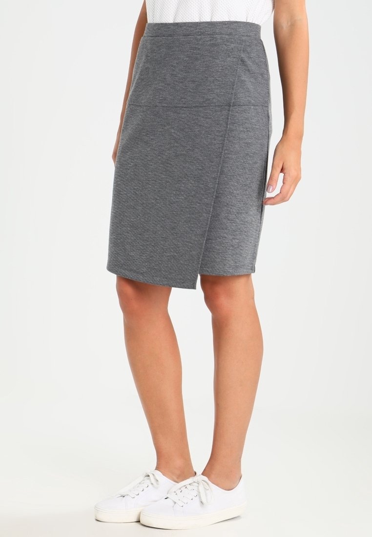 Femme portant une jupe portefeuille grise mi-longue, des baskets blanches et un top blanc sans manches, debout sur un fond clair uni.