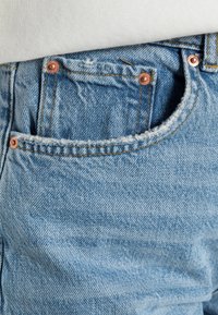 Ljusblå denimjeans med en urblekt struktur, fransiga fickkanter och kopparfärgade knappar. Sömnadsdetaljer synliga längs sömmarna.