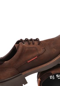 Zapatos de cuero marrón con una textura suave, punta redonda y seis ojales. Presenta un acento rojo cerca de los cordones y una suela texturizada.