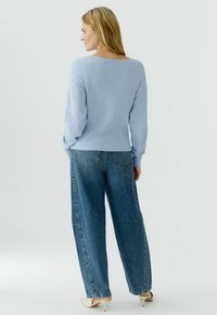 Frau trägt einen hellblauen Pullover mit langen Ärmeln, weite blaue Jeans und weiße High-Heels, steht und schaut nach hinten.