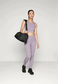 Vaaleanlila lyhyt urheilupaita ja siihen sopivat leggingsit, yhdistettynä mustaan matkakassiin. Mallilla on jalassaan mustat urheilukengät.