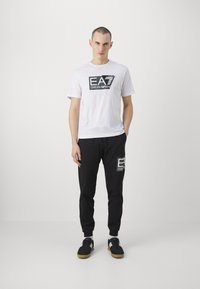 White cotton T-shirt with black graphic logo; black sweatpants with logo on left thigh; worn with black sneakers and white socks.