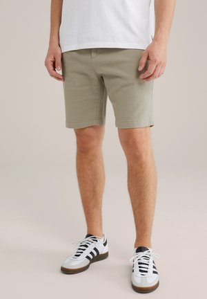 Mann trägt hellbraune, strukturierte Shorts, weißes T-Shirt und weiße Sneakers mit schwarzen Streifen und steht vor einem neutralen Hintergrund.
