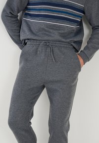 Sudadera gris con rayas azules y blancas en el pecho, combinada con pantalones de chándal grises a juego que tienen cintura con cordón y bolsillos laterales.