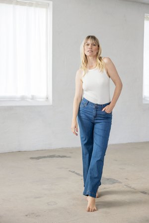 Blonde Frau barfuß in weißem ärmellosem Oberteil und blauen Jeans steht in einem hellen, leeren Raum mit Betonboden und großen Fenstern.
