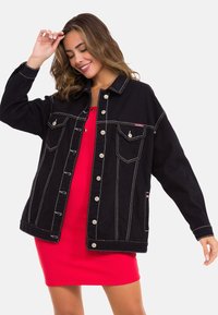 Schwarze Denimjacke mit silbernen Knöpfen, weißen Nähten und zwei Brusttaschen; getragen über einem roten gerippten Kleid.