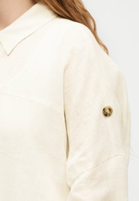 Γυναίκα που φοράει κρεμ μπλούζα με μακριά μανίκια, μυτερό γιακά και διακοσμητικό καφέ κουμπί στο πάνω μέρος του μανικιού.