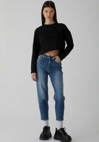 Crna skraćena dukserica s utisnutim tekstom, uparena s plavim farmerkama ravnog kroja i crnim tenisicama preko bijelih čarapa. Jednostavan, ležeran outfit.