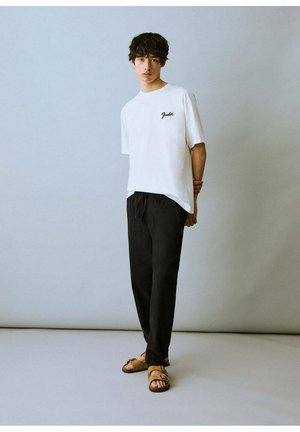 T-shirt blanc à manches courtes avec logo brodé noir, associé à un pantalon noir à cordon de serrage et des sandales marron à plusieurs straps.