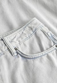 Υφασμα από ανοιχτό μπλε denim που διαθέτει καμπύλη πίσω τσέπη με στρογγυλεμένο σχήμα, λεπτομέρειες με ραφή στο πάνω μέρος και κλείσιμο με μεταλλικό κουμπί.