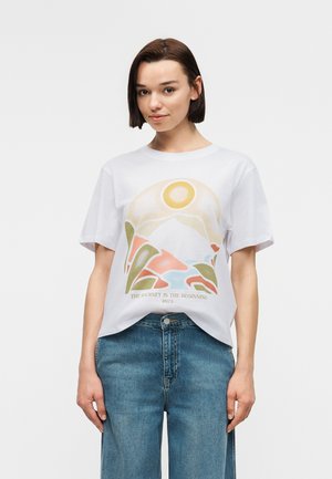 Jeune femme portant un t-shirt blanc avec un motif de soleil, montagne et plantes au-dessus du texte « Le voyage est le commencement » et un jean bleu.