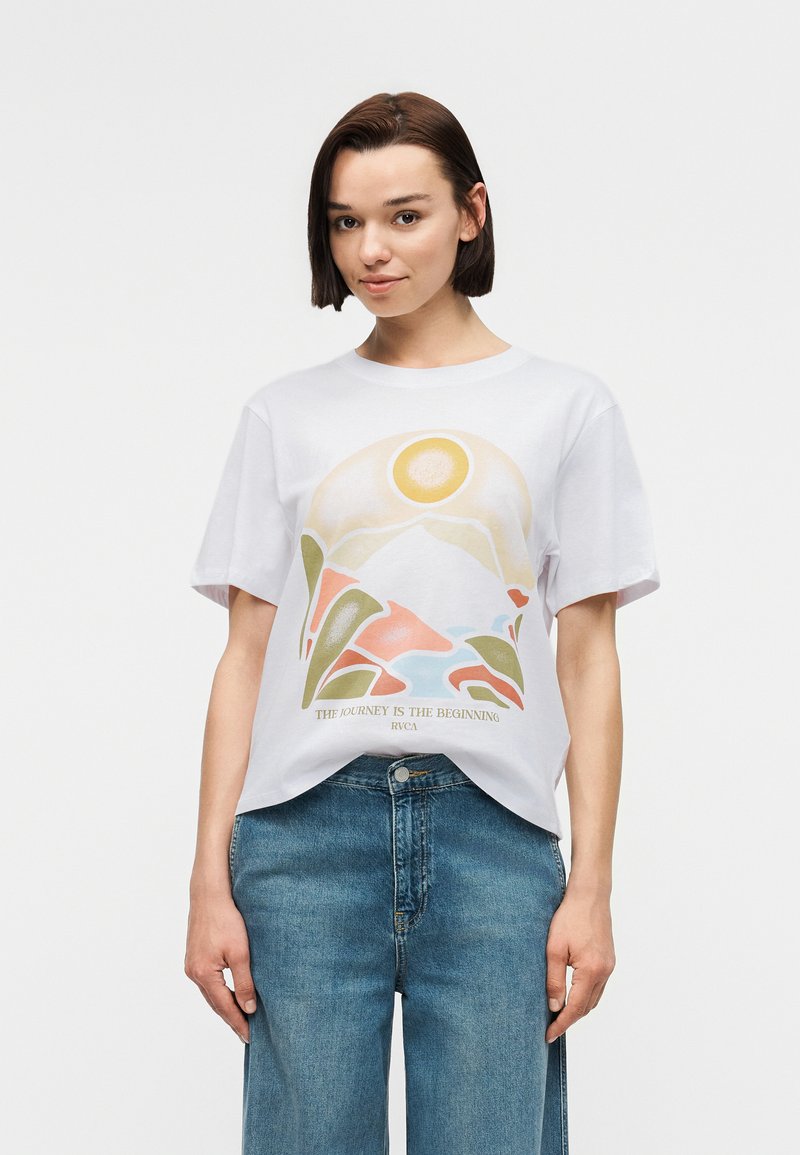 Νεαρή γυναίκα που φοράει λευκό μπλουζάκι με γραφικό ήλιο, βουνό και φυτά πάνω από το κείμενο «Το ταξίδι είναι η αρχή» και μπλε τζιν.