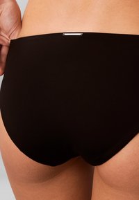 Gros plan sur le bas du dos et les hanches d'une personne portant un sous-vêtement noir Calvin Klein avec une petite étiquette en métal à logo au centre arrière de la ceinture.
