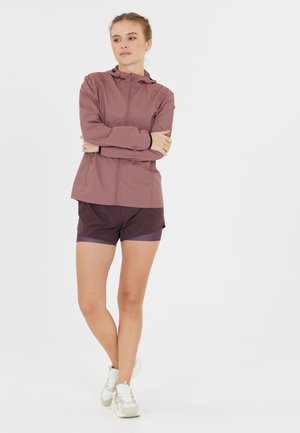Lätt mauve jacka med huva och frontdragkedja; figurnära design. Kombinerad med mörklila shorts och vita sneakers.