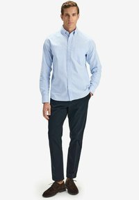 Vaaleansininen raidallinen nappipaita, pitkät hihat, yhdistetty tummiin housuihin ja ruskeisiin mokkanahkakenkiin, seisovalla miesmallilla.