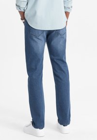 Jeans de denim azul con corte recto, diseño de cinco bolsillos y costuras sutiles. Usados con zapatillas blancas y una camisa de color claro.