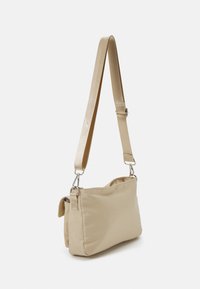 Liten beige axelväska med justerbar rem och silverfästen, något puffat tyg, stående mot en vit bakgrund.