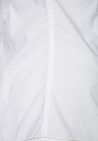 Biały materiał o gładkiej fakturze z widocznym pionowym szwem. Tkanina wydaje się lekka i lekko pomarszczona.