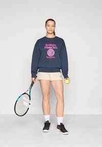Navy sweatshirt with pink logo text, beige shorts, holding a tennis racket and ball, paired with black athletic shoes and white socks.