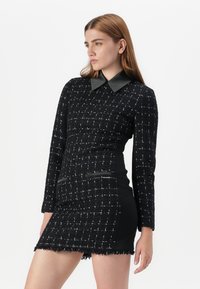 Ein Modell mit langen braunen Haaren posiert in einem eng anliegenden schwarzen Kleid mit strukturiertem Gewebe und einem glänzenden Kragen vor einem weißen Hintergrund.