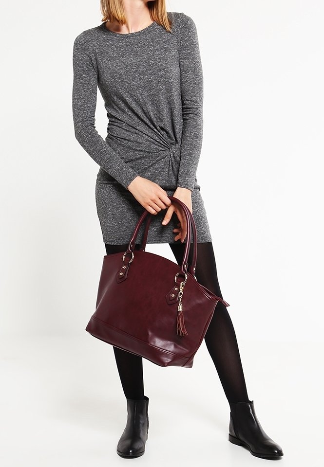 Femme en robe à manches longues grise et collants noirs tenant un grand sac à main bordeaux avec un gland, portant des bottines noires.