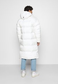 Lungo cappotto bianco imbottito con cappuccio, caratterizzato da un design a confezioni; indossato sopra jeans azzurri chiari e sneakers bianche. Texture liscia e lucida.