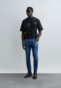 Άνδρας που φοράει μαύρα γυαλιά ηλίου, μαύρο κοντομάνικο με σκούρο κείμενο, στενά μπλε τζιν με φθορές και μαύρα μποτάκια μέχρι τον αστράγαλο, στέκεται μπροστά σε απλή λευκή επιφάνεια.