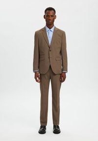 Καφέ κοστούμι με στενή γραμμή, μονό κουμπί, πλαϊνές τσέπες, συνδυασμένο με γαλάζιο πουκάμισο και μαύρα παπούτσια.
