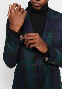 Geruite blazer in donker groen en marineblauw met paarse accenten, met een geknipt kraag, contrasterende zwarte trui met col en knoopmanchetten.