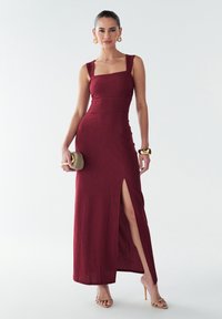 Bordo spalvos megzta maxi suknelė su kvadratiniu iškirpimu, storomis petnešėlėmis, šoniniu skeltuku ir aptemptu siluetu. Priedai – delninė rankinė ir basutės su dirželiais.
