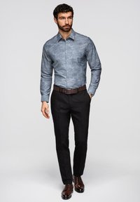 Homme portant une chemise à motifs bleue à boutons, un pantalon noir, une ceinture marron et des chaussures habillées marron, debout avec une main dans la poche.