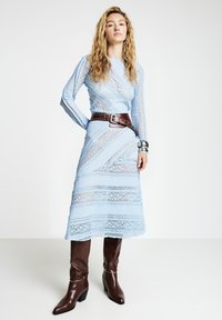 Robe à manches longues en dentelle bleu clair avec une silhouette ajustée, dotée d'une taille ceinturée marron, accompagnée de bottes marron à talons montant jusqu'au genou.