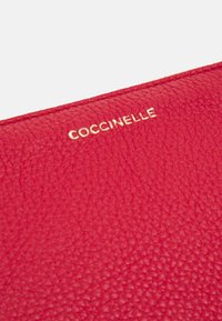 Rødt læderpung med en tekstureret overflade og præget guldlogo "Coccinelle". Kompakt, rektangulært design med rene sømme.