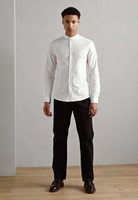 Camisa blanca abotonada con cuello mao, combinada con pantalones negros y zapatos de cuero marrón, frente a un fondo gris claro.