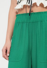 Pantalon vert à jambes larges en tissu texturé, avec une ceinture élastique et des poches latérales. Le tissu a un léger éclat.