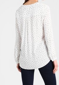 Blusa bianca con un motivo geometrico in nero, rosso e blu. Maniche lunghe con polsini arruffati. Orlo arrotondato e tessuto morbido e leggero.