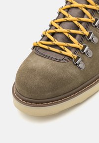 Bota de senderismo de ante marrón con cordones amarillos, ganchos de metal y puntera texturizada. Presenta una suela de goma marrón cosida y una forma redonda.