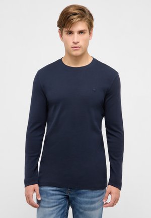 Młody mężczyzna w granatowej koszulce z długim rękawem i okrągłym dekoltem oraz niebieskich dżinsach, stojący na tle jednolitego jasnego tła.