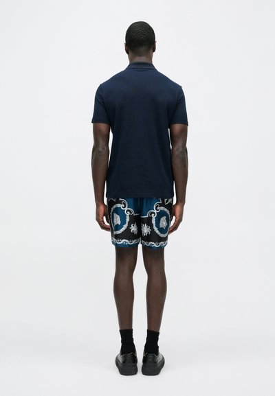 Μπλε ναυτική πόλο μπλούζα συνδυασμένη με σορτς με σχέδιο σε μπλε και μαύρο, περίτεχνα λευκ�ά στοιχεία και μαύρα παπούτσια.