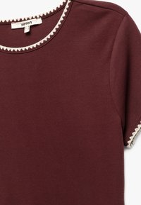 Burgundy t-shirt i mjukt bomull, med rund halsringning och vitt triangulärt mönster samt kortärmar med liknande detaljer.