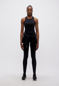 Γυναίκα που στέκεται με το πρόσωπο μπροστά, φορόντας μαύρη αθλητική στολή με φούτερ δεμένο γύρω από τη μέση και μαύρα αθλητικά παπούτσια.