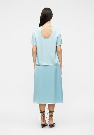 Γυναίκα με ανοιχτό γαλάζιο σατέν τοπ με κοντό μανίκι και άνοιγμα στην πλάτη και αντιστοιχισμένη μίντι φούστα, φορώντας μαύρα δερμάτινα παπούτσια με κορδόνια, στέκεται με την πλάτη στραμμένη.