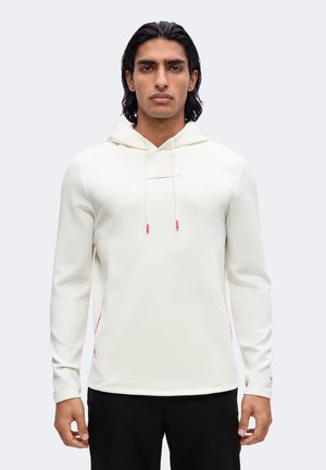 Mann mit langen dunklen Haaren trägt ein weißes Kapuzensweatshirt mit rosa Kordelenden und schwarze Hosen, stehend vor einem schlichten Hintergrund.