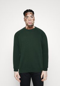 Tmavě zelená mikina s kulatým výstřihem a raglánovými rukávy, vyrobená z měkké látky. Má žebrované manžety a lem. Nošena s černými kalhotami.