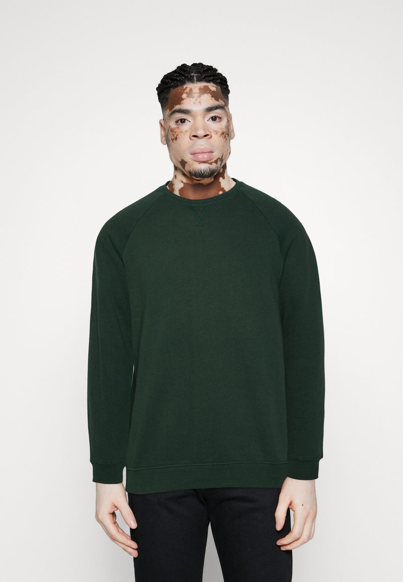 Tmavě zelená mikina s kulatým výstřihem a raglánovými rukávy, vyrobená z měkké látky. Má žebrované manžety a lem. Nošena s černými kalhotami.